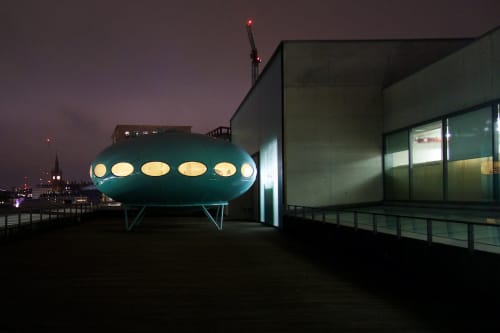 Facing straight on Futuro House. It’s on the roof of a building on a cloudy night.