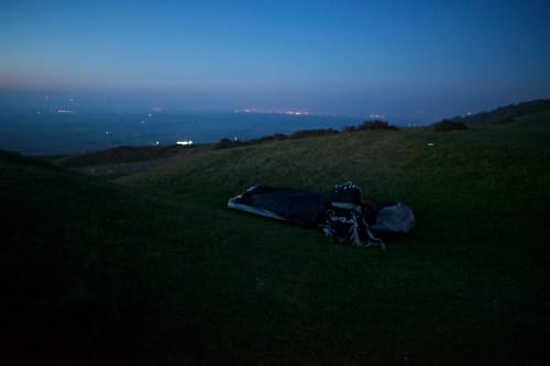 A bivvy bag and rucksack sit in a hollow depression in the grass in the last of the evening light. The sky is dark blue and calm.