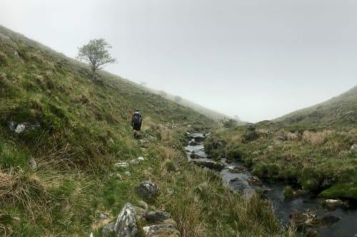 A panorama taking on a foggy day from the side of a small stream. A hiker is ahead on the stream walking downhill.