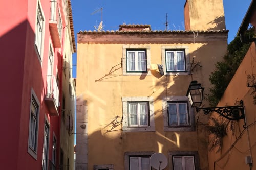A portrait photo of the entrance to a 4 storey building at the end of a wide alleyway. The building is painted in pastel-yellow, and has a pastel pink building to the left.