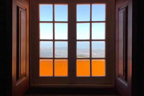 Looking from inside to a window looking out on a high vista. The interior is dark with dark wood window frame. Through the window the sky outside is bright. The window is made from two sets of 2x4 small glass frames. The bottom row is bright orange from an balcony outside.