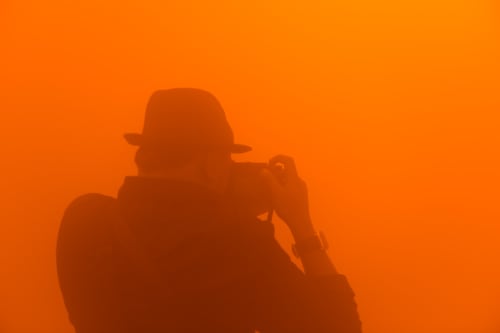 A photographer wearing a hat composes a photo inside an orange foggy room.