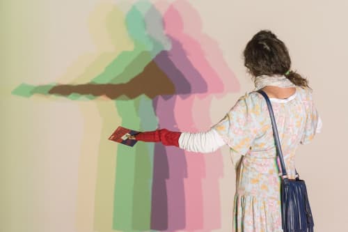 A woman stands in front of a white wall. A light shines on her from behind, casting a multi-coloured shadow on the wall.
