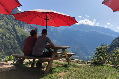 Ed and Oj sat at a picnic bench half way up a valley. There’s a large red awning above them.