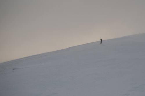 A wide shot at dusk looking up a snowy hill. A figure (Ed) in the distance can be seen waving an ice axe.