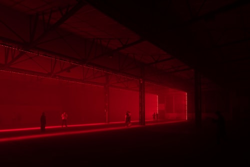 A photo in a large dark warehouse. The only light is deep red in one section of the warehouse. Several still figures can be seen around.