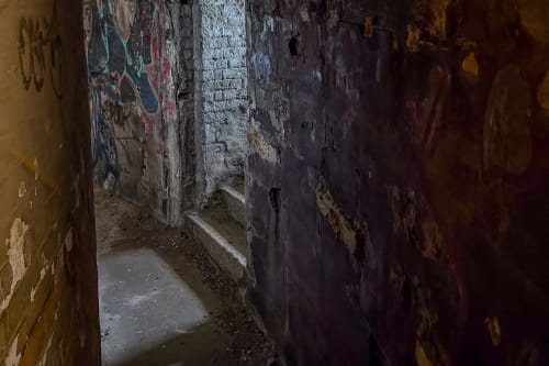 Dark photo taken in a curving corridor. The only light comes from an exit going to the right and up some stairs. Graffiti can just be made out on the walls.
