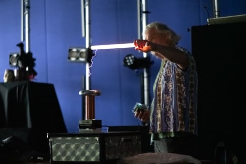 Man in a dark room holding a 2′ fluorescent tube in front of a small tesla coil. The tube is lit up and there are purple sparks from the coil to the tube.