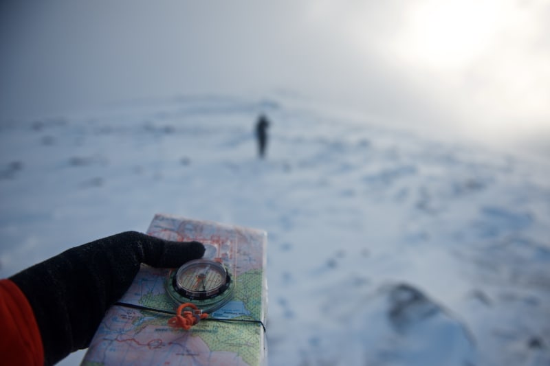 A hand in the foreground holds a compass and map. In the background is a figure walking away. The compass is pointing towards the figure.