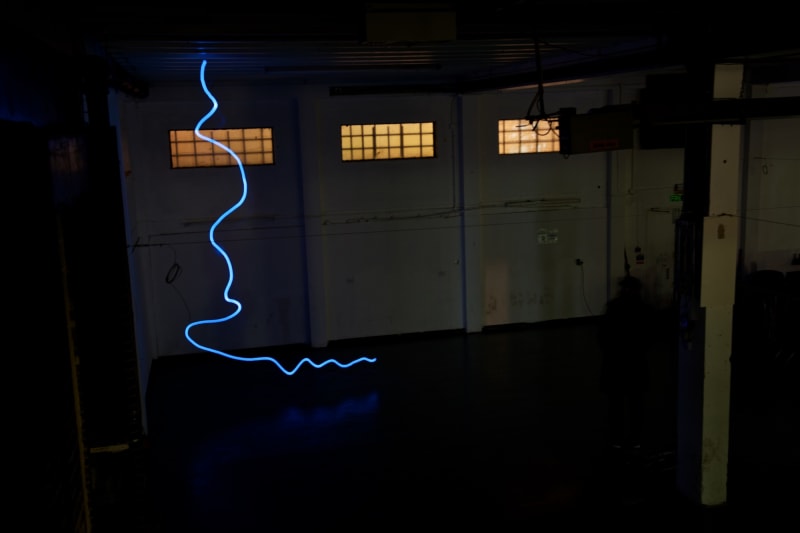 A photo taken in a dark warehouse from above, looking down. There’s a single blue line of light snaking from the floor to the ceiling.