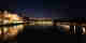 A panorama taken from a bridge over the Rhine at nighttime. There are buildings in the distance lit up and lights reflected in the water.