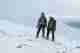 Ed and Chris pose together facing the camera on the edge of Aonach Mòr. Both are wearing green jackets and are tied together with a green climbing rope.