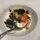 A close up of a bowl of food. The bowl contains a single scallop in a white cream sauce. There’s scattered fried seaweed on top.