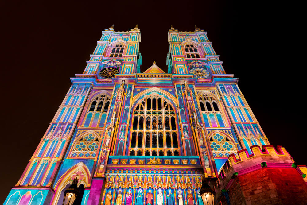 A photo of the front of Westminster Abbey at night. The entire front is lit up in bright pastel colours.