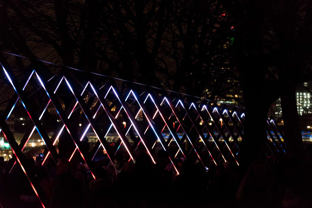 A photo of a light triangular light sculpture. A series of triangles is each lit up in white and bright colours. The triangles recede in to the distance.