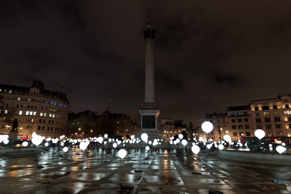 Many balloons float above the ground of Trafalgar Square - the balloons are lit like light bulbs.