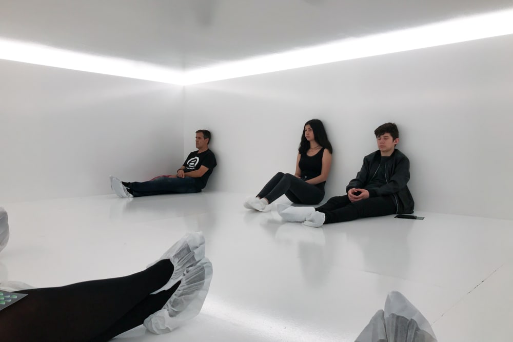 Several people with paper socks are sat in a gloss white room with white light overhead.