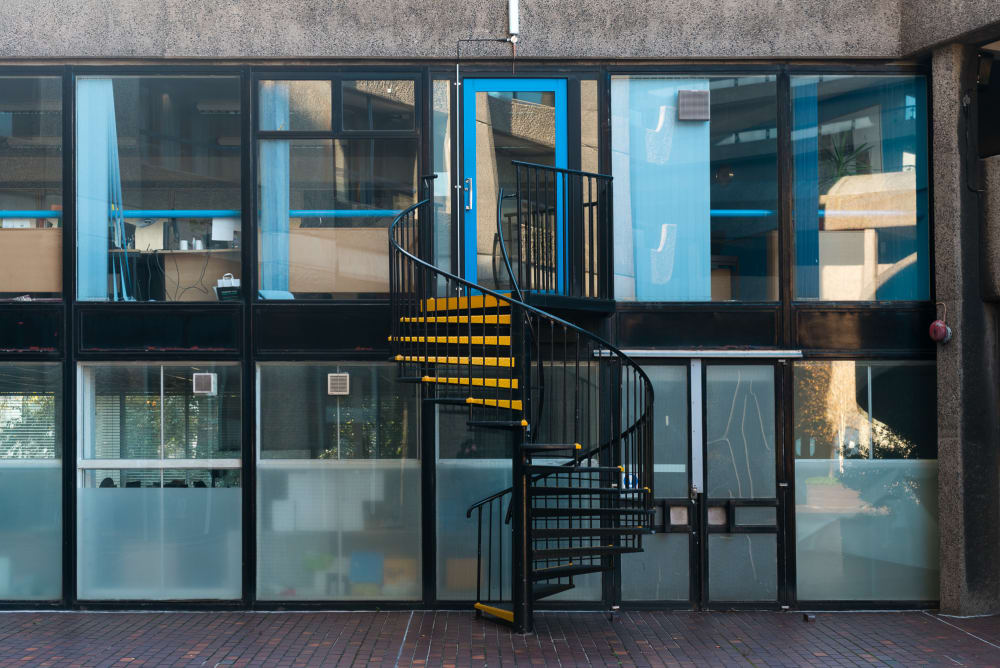 Facing the exterior of a glass fronted building straight on. There’s a metal spiral staircase with yellow treads connecting the two floors.