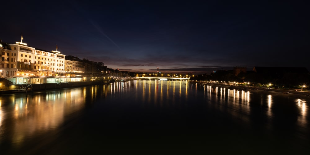 A panorama taken from a bridge over the Rhine at nighttime. There are buildings in the distance lit up and lights reflected in the water.