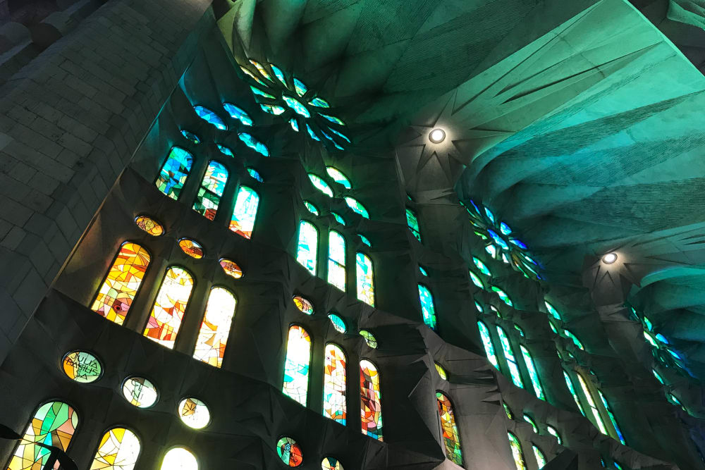 Looking up at some stained glass windows in la Sagrada Família. The near ceiling is faceted and coloured green and blue from the window light.