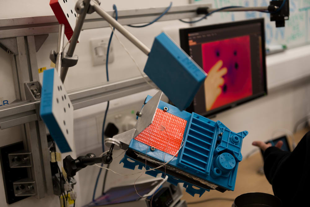 A photo of a science experiment. There are three blue stands held in the air. There’s a screen in the background with a hand visible on it.