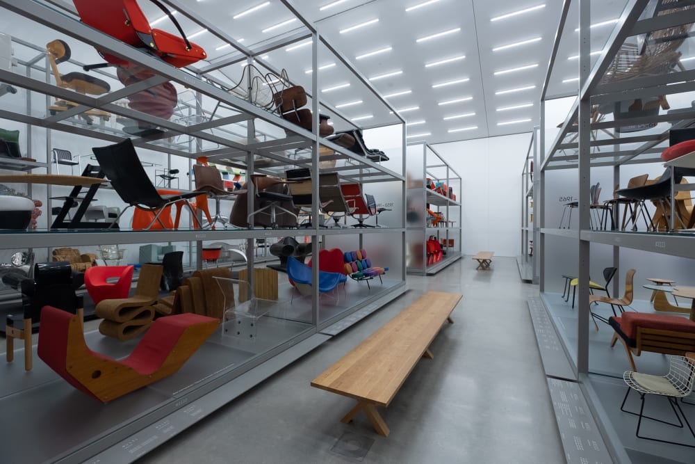 Inside the Vitra Design Museum archive. The room is brightly lit with aluminium shelves on each side receding in to the distance. On the shelves are many different chairs.