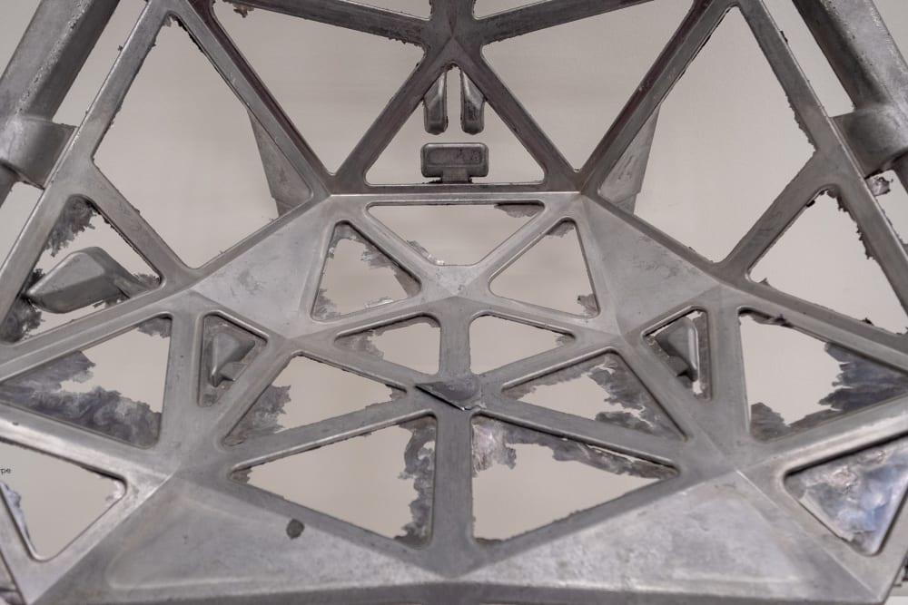 A detail shot of an unfinished metallic form of a Konstantin Grcic chair. The form is made of metal struts connected together with waste metal from the mould still visible.