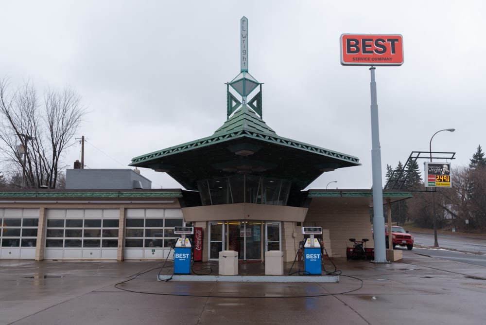 A photo of the front of the Frank Lloyd Wright gas station. There’s gas pumps in the front and a tall sign reading ‘BEST’.