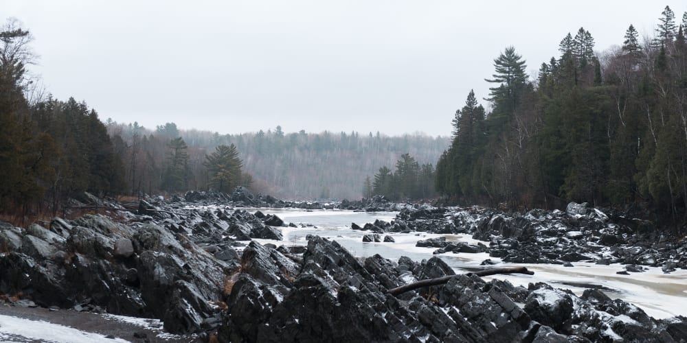 A long view across an icy river. There’s dark rocky outcrops coming out of the river. The river is partially frozen with creamy patches of ice at the sides.