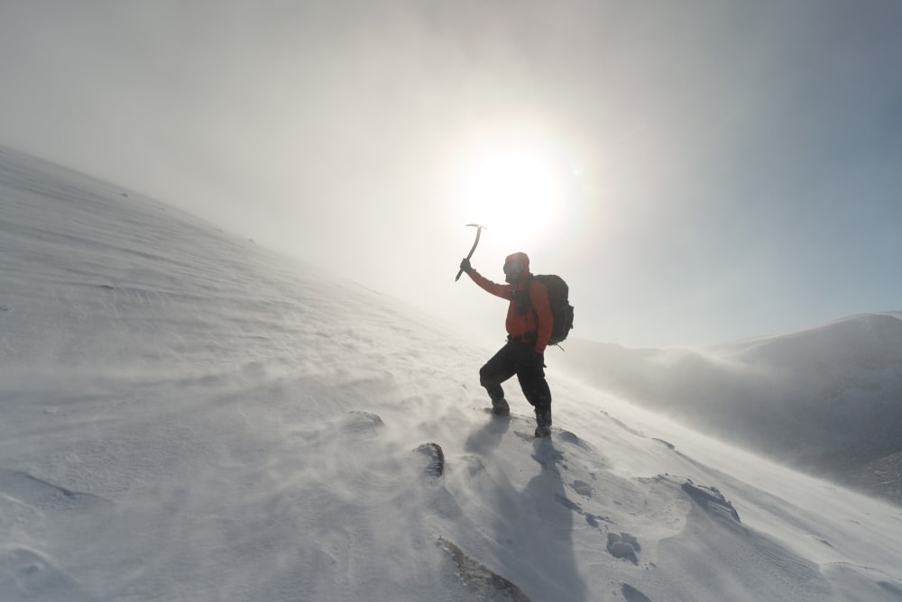 Photo looking up a snowy hillside. Ed is in the foreground holding an axe above his head. The sun is nearly directly behind him.