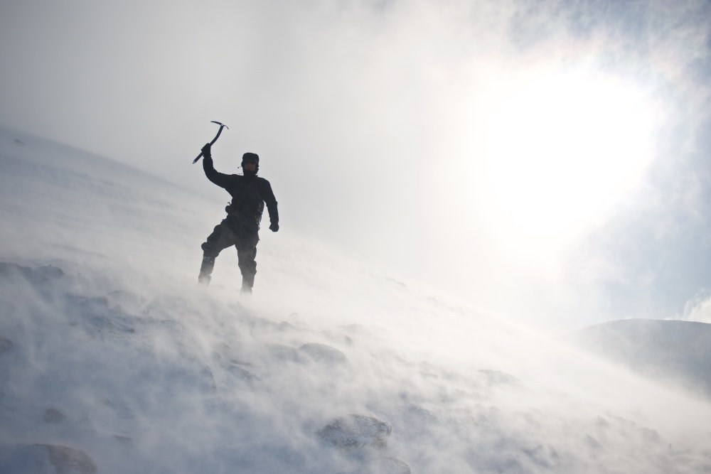 A photo looking up a snowy mountain - in the near distance is Chris Natt holding an ice axe over his head. There’s flurries of snow all around.