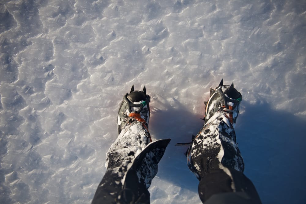 Looking down at a pair of boots with crampons on icy ground.