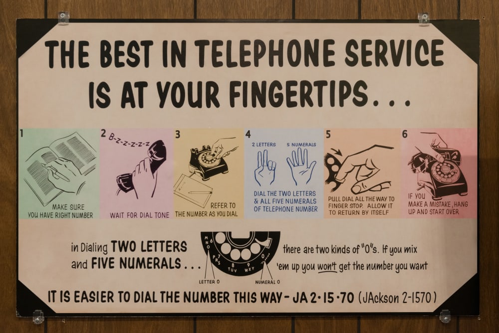 Photo of a vintage poster describing with words and diagrams how to use a rotary telephone. The main reading reads “The best in telephone service is at your fingertips...” with a 6 step diagram for how to use a rotary phone.