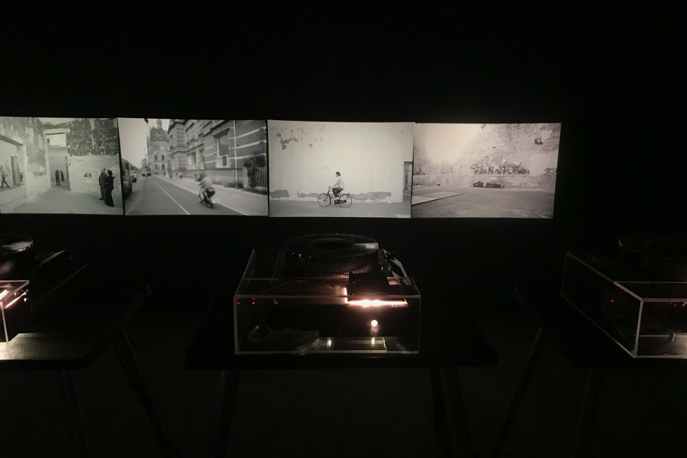 Five projectors in a dark room project black and white photos on to a wall.