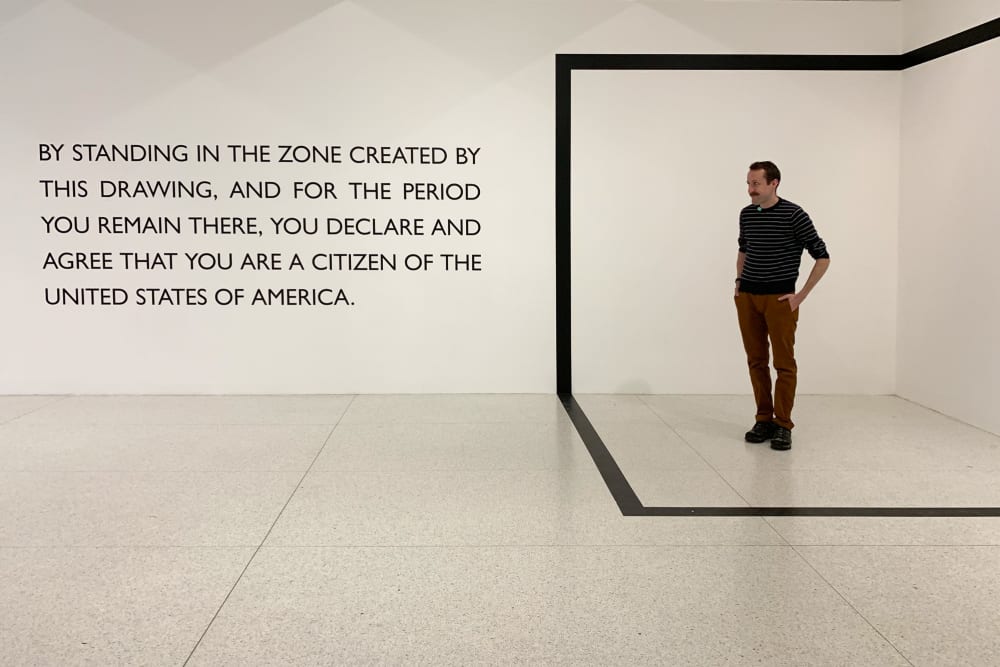 An area is marked off in the corner of a room in the shape of a cube. Ed Horsford stands in the area. To the left is a sign that reads “By standing in the zone created by this drawing, and for the period you remain there, you declare and agree that you are a citizen of the United States of America.”.