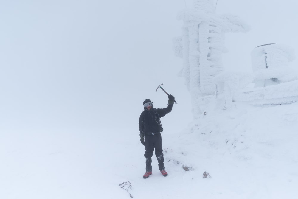 Ed Horsford standing next to the Cairngorm weather station, holding an ice axe over his head. The weather station is completely iced over and there’s very little visibility.