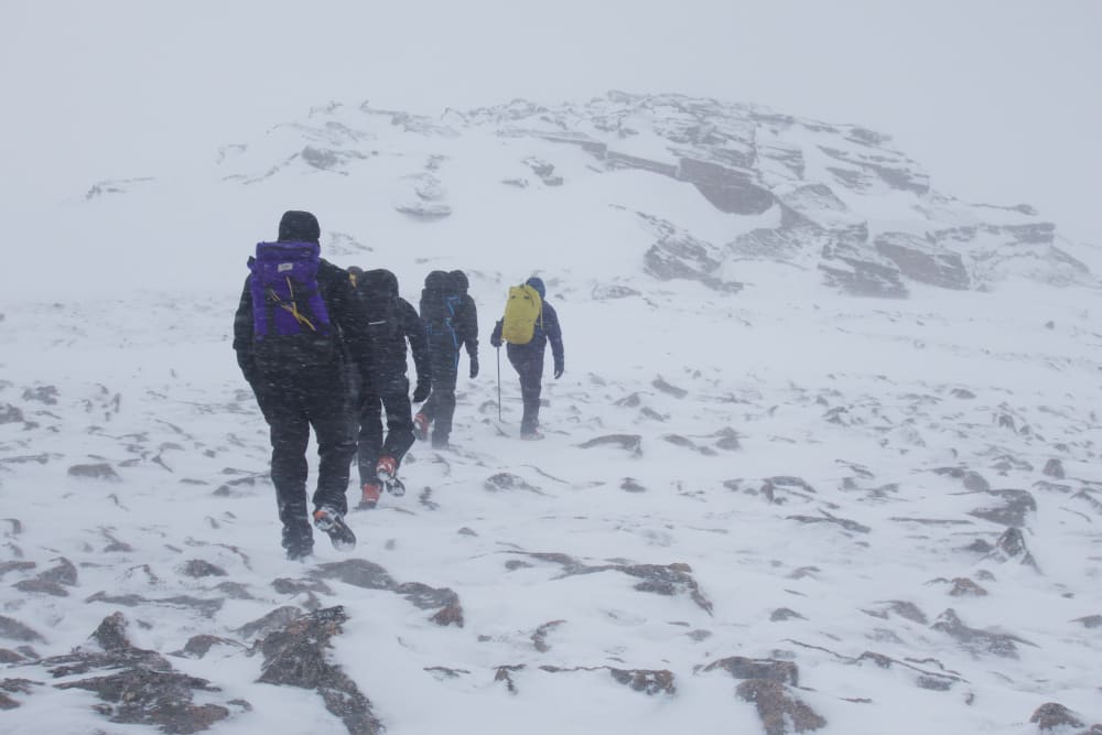 Four men dressed in winter gear walk in a line up a snowy mountain in poor visibility.