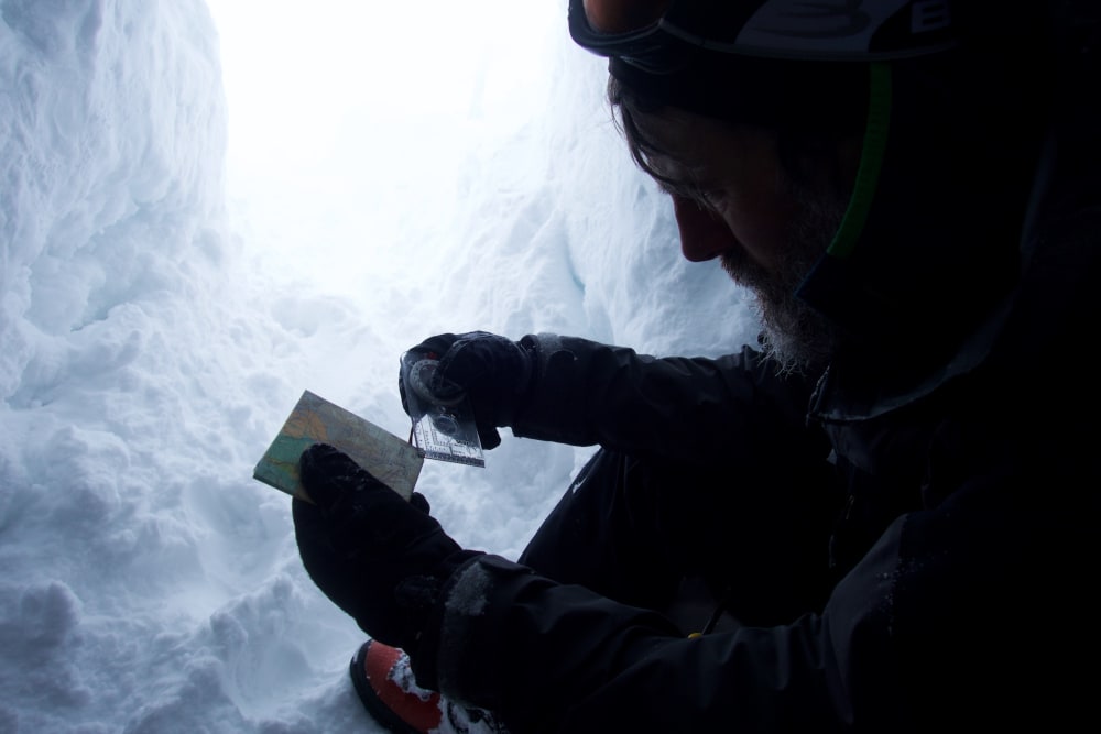 A man uses the corner of a compass to look at a map. He’s sat inside a snow hole.