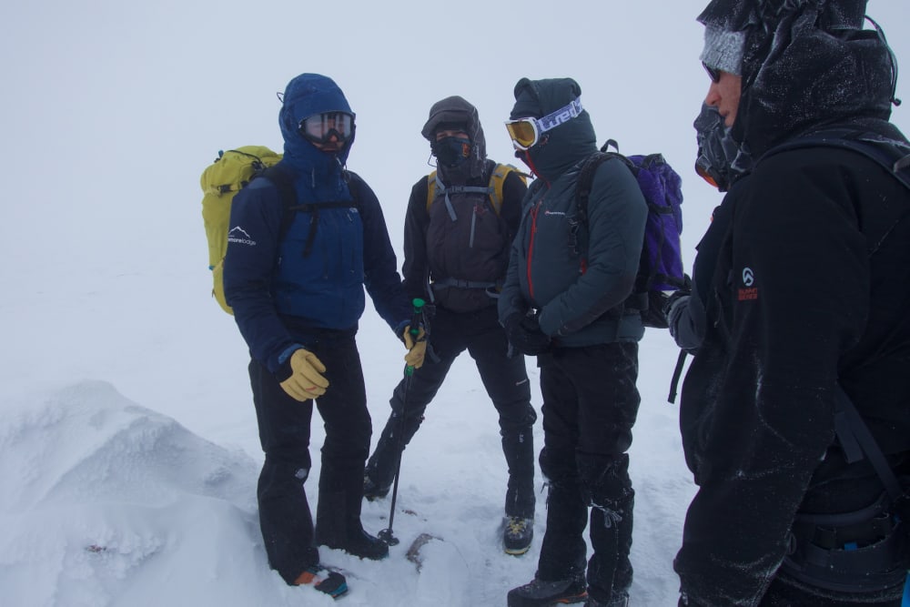 Four men wrapped up in winter clothing huddle on a snowy mountain.