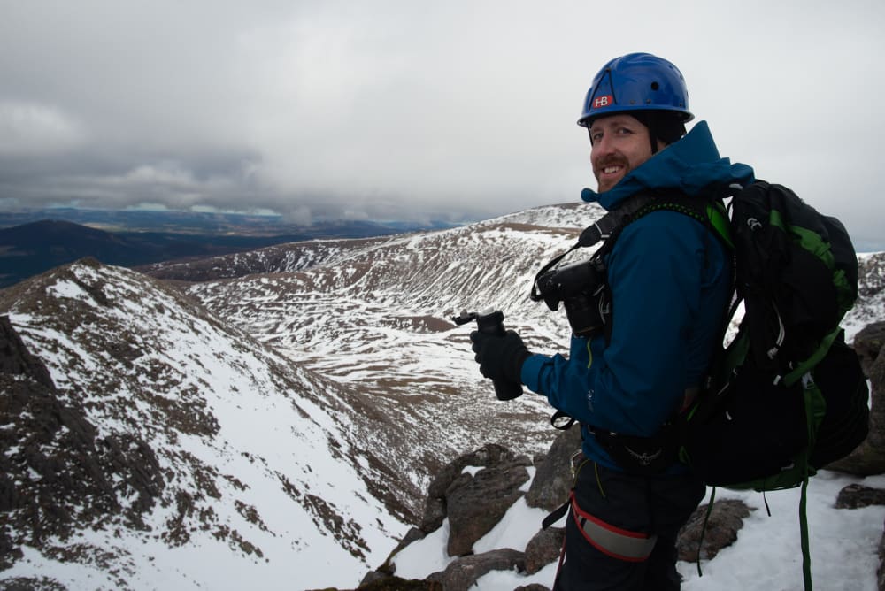 Ed drinking from a vacuum flask, with snowy mountainside in the distance. He’s wearing full climbing gear - helmet, harness, rucksack.