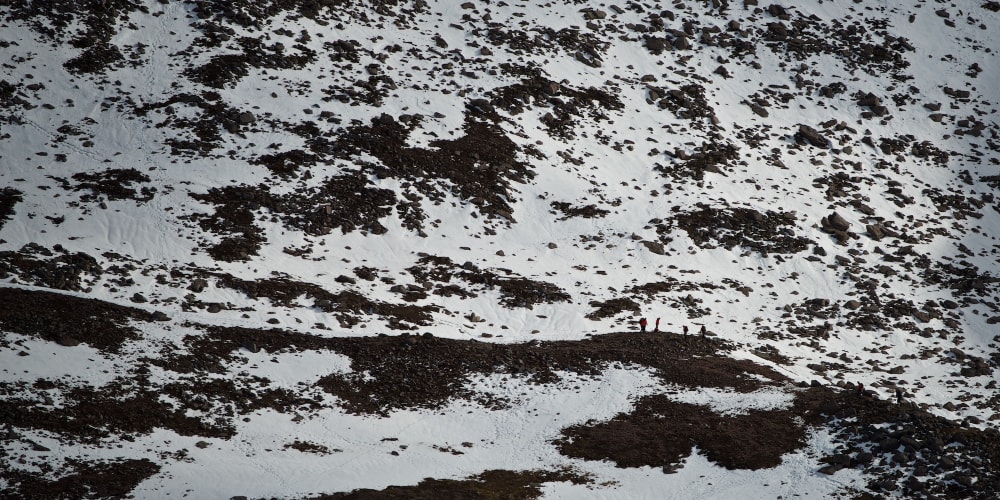 A long photo of several hikers in the distance walking along a snowy path on a mountain side.