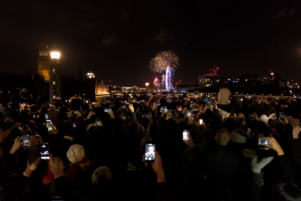 A crowd on Lambeth Bridge watch the New Years fireworks display over the London Eye. Many are holding phones in the air taking photos.