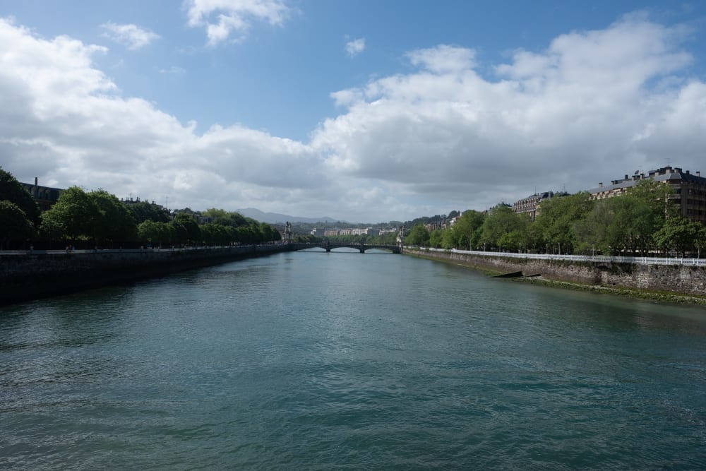 Looking directly down the Urumea river to the next bridge. There’s lots of green trees on each side of the bank, and the water is calm.