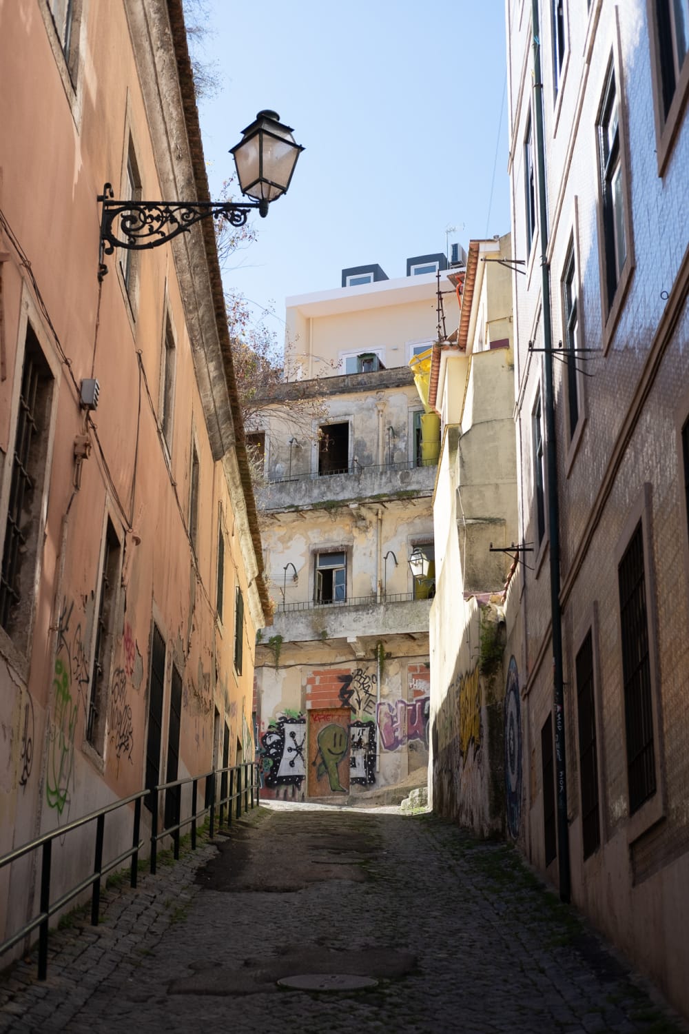 Looking up an alleyway. The foreground is dark and in shadow, but the background is brightly lit by sun. The alley ends with a light coloured building covered in graffiti.