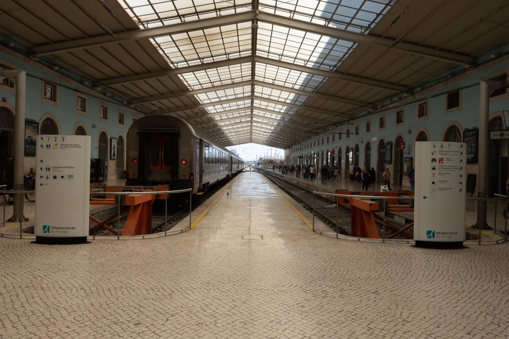 Looking directly down the central platform in a train station. On the left a train is waiting. The station is small and with a low roof.