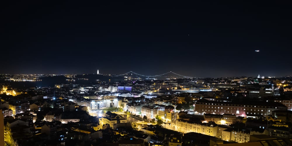 A panorama of Lisbon at night. The streets are filled with yellow and white lights and a suspension bridge can be seen in the distance.