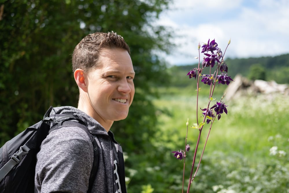 Oj smiling as he looks at the camera and holds some purple flowers.