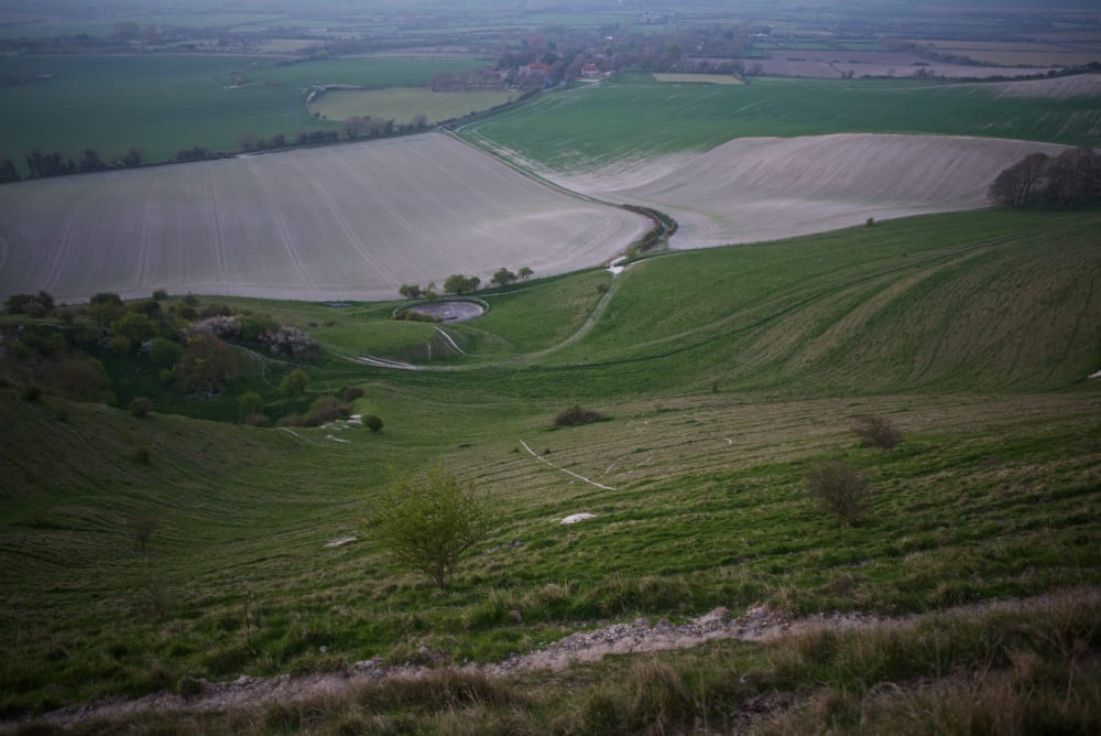 Looking down a grassy hill with a farm in the distance. A faint chalky line can be seen on the ground - part of the Long Man of Wilmington.