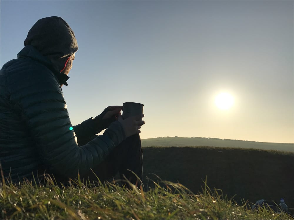 A photo at sunrise of Ed Horsford sitting on a grassy hill with a cup in hand looking over a hill.