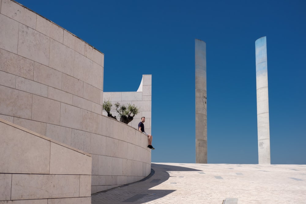 A photo taken in the courtyard of the Champalimaud Foundation. There’s a raised platform on the left, with Ed Horsford sat looking at the camera. On the right are two tall concrete towers set against a very deep blue sky.